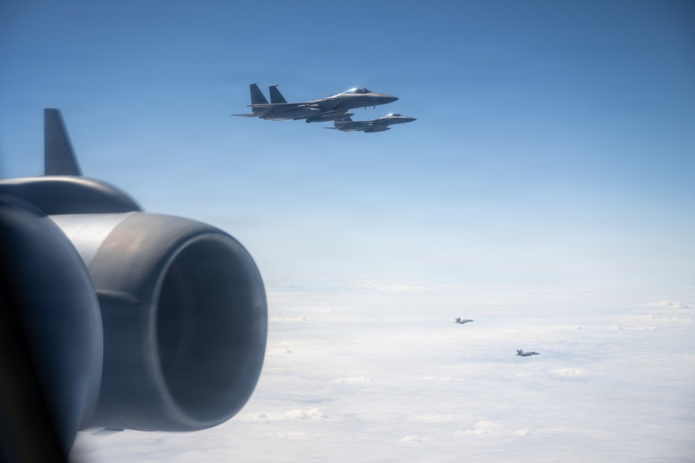 An aerial photograph taken from inside or alongside a large aircraft showing four military fighter jets in formation flight above a cloud layer. Two jets are clearly visible in close proximity in the upper portion of the frame, while two additional jets are visible at greater distance in the background. The foreground shows what appears to be a jet engine nacelle or exhaust nozzle from the camera platform aircraft.