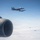 An aerial photograph taken from inside or alongside a large aircraft showing four military fighter jets in formation flight above a cloud layer. Two jets are clearly visible in close proximity in the upper portion of the frame, while two additional jets are visible at greater distance in the background. The foreground shows what appears to be a jet engine nacelle or exhaust nozzle from the camera platform aircraft.