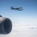 An aerial photograph taken from inside or alongside a large aircraft showing four military fighter jets in formation flight above a cloud layer. Two jets are clearly visible in close proximity in the upper portion of the frame, while two additional jets are visible at greater distance in the background. The foreground shows what appears to be a jet engine nacelle or exhaust nozzle from the camera platform aircraft.