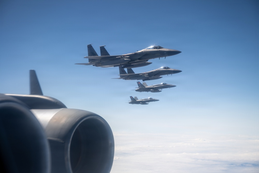 A formation of four military jet aircraft flying in a diagonal echelon formation against a clear blue sky. The photo is taken from another aircraft, with a jet engine nacelle visible in the foreground lower-left. The formation appears to consist of mixed aircraft types, including what appear to be F-15 Eagles and F/A-18 Hornets.