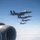A formation of four military jet aircraft flying in a diagonal echelon formation against a clear blue sky. The photo is taken from another aircraft, with a jet engine nacelle visible in the foreground lower-left. The formation appears to consist of mixed aircraft types, including what appear to be F-15 Eagles and F/A-18 Hornets.