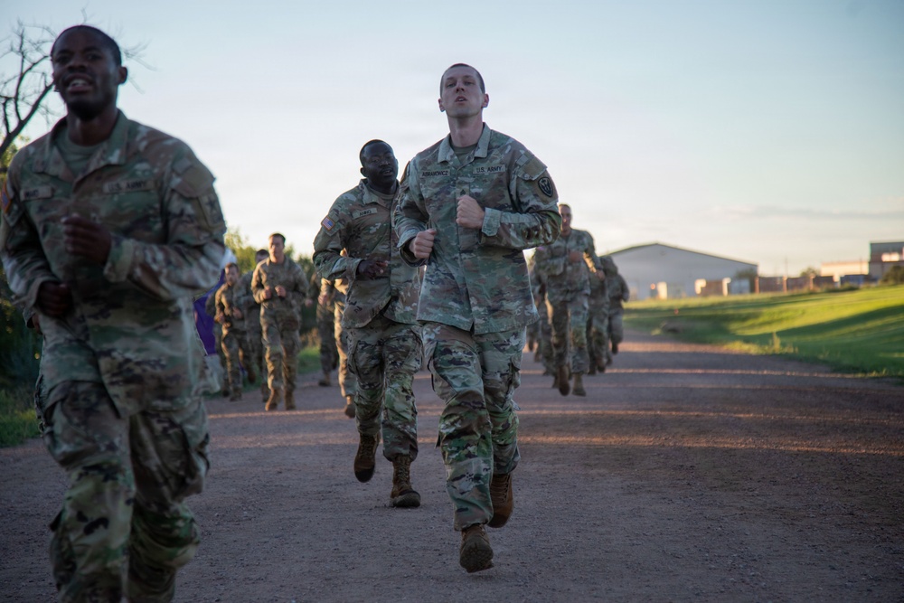 A group of U.S. Army soldiers in OCP (Operational Camouflage Pattern) uniforms are conducting a formation run along a gravel path, likely during physical training (PT). The soldiers are running in a group formation with a building and grassy area visible in the background. The image appears to be taken during early morning hours based on the lighting conditions.