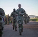 A group of U.S. Army soldiers in OCP (Operational Camouflage Pattern) uniforms are conducting a formation run along a gravel path, likely during physical training (PT). The soldiers are running in a group formation with a building and grassy area visible in the background. The image appears to be taken during early morning hours based on the lighting conditions.