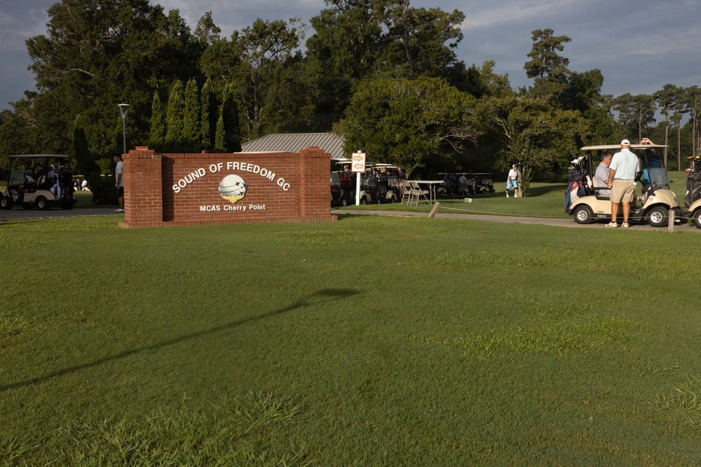 The image shows the entrance sign for 'Sound of Freedom GC' (Golf Course) at MCAS Cherry Point, a Marine Corps Air Station. Multiple golf carts and players are gathered in what appears to be a tournament or group outing staging area. The scene is set on a well-maintained golf course with lush green fairways and mature trees in the background.