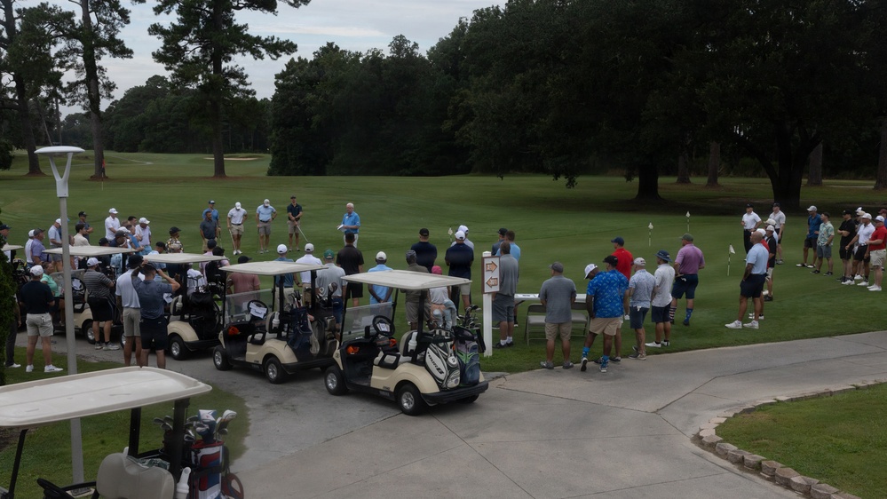 A large group of golfers has gathered at what appears to be a golf course staging area or first tee, likely for a tournament or scramble event. Multiple golf carts are lined up on a cart path while participants stand in clusters on and around the fairway. The scene suggests a pre-round briefing or shotgun start preparation.
