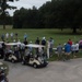 A large group of golfers has gathered at what appears to be a golf course staging area or first tee, likely for a tournament or scramble event. Multiple golf carts are lined up on a cart path while participants stand in clusters on and around the fairway. The scene suggests a pre-round briefing or shotgun start preparation.