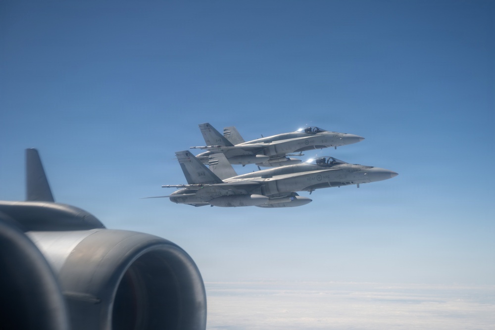 Two military jet fighters, identifiable as F/A-18 Hornet variants, are flying in close formation against a clear blue sky. The photo is taken from aboard a larger aircraft, with a jet engine nacelle visible in the foreground left. The fighters appear to be on an escort or formation flight mission.
