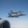Two military jet fighters, identifiable as F/A-18 Hornet variants, are flying in close formation against a clear blue sky. The photo is taken from aboard a larger aircraft, with a jet engine nacelle visible in the foreground left. The fighters appear to be on an escort or formation flight mission.