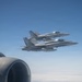 Two military jet fighters, identifiable as F/A-18 Hornet variants, are flying in close formation against a clear blue sky. The photo is taken from aboard a larger aircraft, with a jet engine nacelle visible in the foreground left. The fighters appear to be on an escort or formation flight mission.