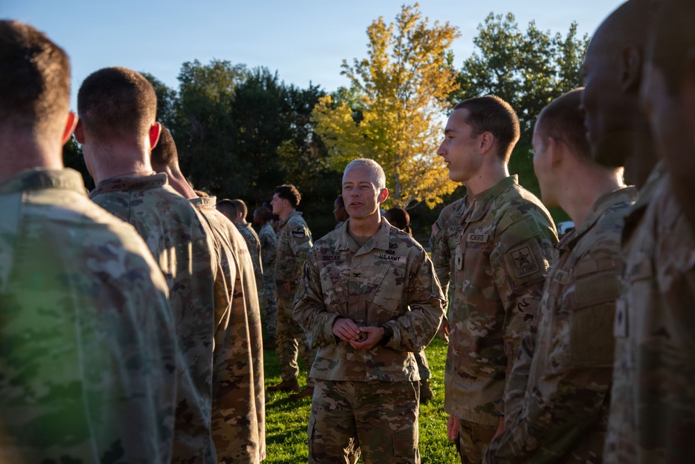 A group of U.S. Army soldiers in OCP (Operational Camouflage Pattern) uniforms are gathered outdoors in what appears to be a briefing or informal discussion. A central figure, identifiable as a higher-ranking officer by visible rank insignia (appears to be a Colonel based on eagle device) and 'U.S. ARMY' tape, is addressing the assembled soldiers. The setting is a grassy outdoor area with autumn-colored trees in the background.