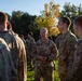 A group of U.S. Army soldiers in OCP (Operational Camouflage Pattern) uniforms are gathered outdoors in what appears to be a briefing or informal discussion. A central figure, identifiable as a higher-ranking officer by visible rank insignia (appears to be a Colonel based on eagle device) and 'U.S. ARMY' tape, is addressing the assembled soldiers. The setting is a grassy outdoor area with autumn-colored trees in the background.
