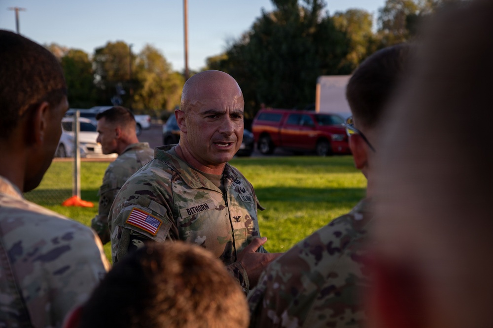 A U.S. Army Colonel named Bithorn, identifiable by the eagle insignia on his uniform, is engaged in conversation with a group of soldiers in an outdoor setting. The scene appears to take place during early morning or late afternoon based on the warm, low-angle lighting. Multiple personnel in OCP (Operational Camouflage Pattern) uniforms are gathered in what appears to be a briefing or informal discussion.