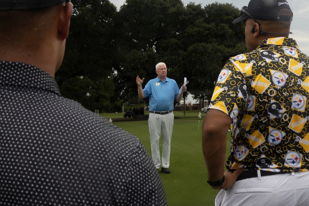 An older white-haired man in a light blue polo shirt and white pants appears to be addressing a small group on a golf course green, gesturing with one hand while holding papers in the other. Two men are visible from behind in the foreground — one wearing a black dotted shirt and another wearing a Pittsburgh Steelers-themed black and yellow patterned shirt. The setting appears to be an organized golf event or tournament briefing.