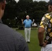 An older white-haired man in a light blue polo shirt and white pants appears to be addressing a small group on a golf course green, gesturing with one hand while holding papers in the other. Two men are visible from behind in the foreground — one wearing a black dotted shirt and another wearing a Pittsburgh Steelers-themed black and yellow patterned shirt. The setting appears to be an organized golf event or tournament briefing.