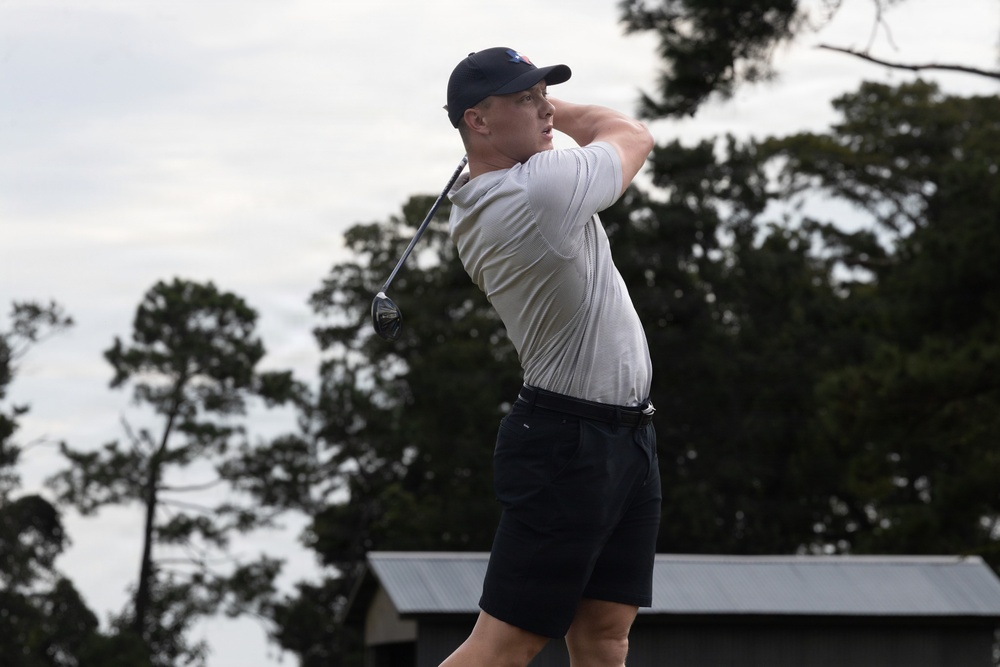 A male golfer is captured mid-swing in a follow-through position, suggesting he has just completed a drive shot. He is dressed in casual golf attire and is photographed against a backdrop of trees and an overcast sky. The image appears to be taken at a golf course during daytime.