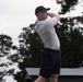 A male golfer is captured mid-swing in a follow-through position, suggesting he has just completed a drive shot. He is dressed in casual golf attire and is photographed against a backdrop of trees and an overcast sky. The image appears to be taken at a golf course during daytime.