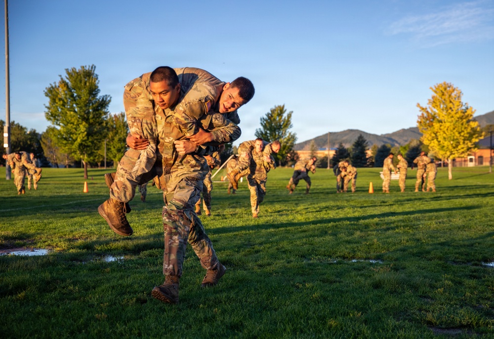 Military personnel in OCP (Operational Camouflage Pattern) uniforms are conducting a physical training exercise on a grass field during early morning or late afternoon, as indicated by the warm golden light. The foreground shows one soldier carrying another on his back in what appears to be a buddy carry drill, while multiple pairs of soldiers perform the same exercise in the background. The setting appears to be a military installation with mountains and trees visible in the distance.