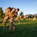 Military personnel in OCP (Operational Camouflage Pattern) uniforms are conducting a physical training exercise on a grass field during early morning or late afternoon, as indicated by the warm golden light. The foreground shows one soldier carrying another on his back in what appears to be a buddy carry drill, while multiple pairs of soldiers perform the same exercise in the background. The setting appears to be a military installation with mountains and trees visible in the distance.