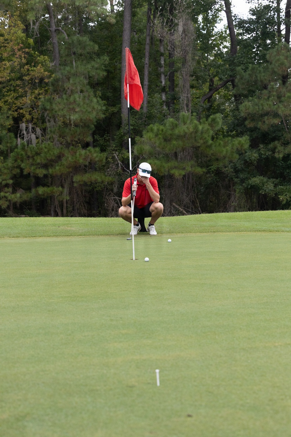 A golfer dressed in a red shirt, black shorts, and white cap is crouching on a putting green near the flagstick, appearing to read the green and assess his putt. Two golf balls are visible on the green, suggesting a practice or competitive putting situation. The scene is set on a well-maintained golf course surrounded by dense trees in the background.