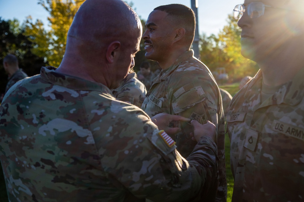 A U.S. Army ceremony is taking place outdoors where a senior soldier is pinning something onto the uniform of a smiling soldier, likely a promotion or award ceremony. The recipient appears pleased and is smiling broadly while the presenter works on his chest area. A third soldier in U.S. Army uniform is visible on the right side of the frame.