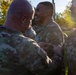 A U.S. Army ceremony is taking place outdoors where a senior soldier is pinning something onto the uniform of a smiling soldier, likely a promotion or award ceremony. The recipient appears pleased and is smiling broadly while the presenter works on his chest area. A third soldier in U.S. Army uniform is visible on the right side of the frame.
