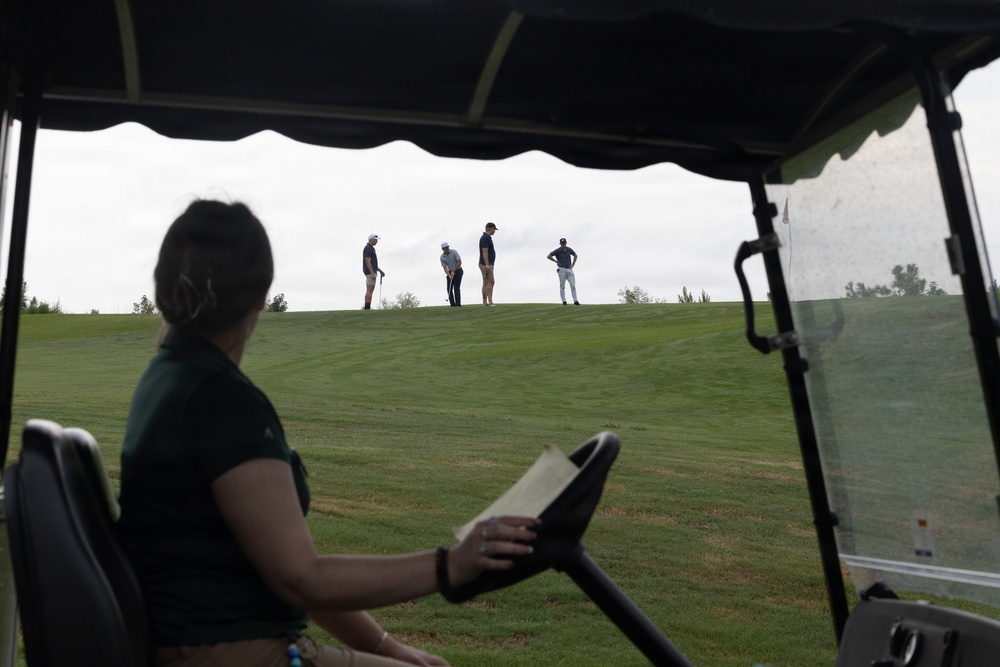 The image is taken from inside a golf cart, showing a person in the foreground seated at the wheel holding what appears to be a scorecard or document. In the background, four golfers are visible standing on an elevated green or fairway ridge against an overcast sky. The perspective creates a layered composition with the cart interior framing the distant players.