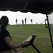 The image is taken from inside a golf cart, showing a person in the foreground seated at the wheel holding what appears to be a scorecard or document. In the background, four golfers are visible standing on an elevated green or fairway ridge against an overcast sky. The perspective creates a layered composition with the cart interior framing the distant players.