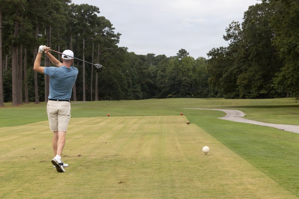 A male golfer is captured mid-swing on a golf course tee box, having just completed a drive shot. The fairway stretches ahead lined with tall pine and deciduous trees on both sides, with a cart path visible on the right. Multiple colored marker balls are visible along the fairway, likely indicating distance markers.