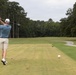A male golfer is captured mid-swing on a golf course tee box, having just completed a drive shot. The fairway stretches ahead lined with tall pine and deciduous trees on both sides, with a cart path visible on the right. Multiple colored marker balls are visible along the fairway, likely indicating distance markers.