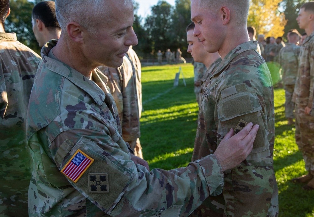 A senior U.S. Army soldier is pinning a unit patch onto the sleeve of a younger soldier during what appears to be a formal military ceremony outdoors. Both soldiers are wearing OCP (Operational Camouflage Pattern) uniforms, and the senior soldier has a 4th Infantry Division patch and U.S. flag on his left sleeve. Multiple other soldiers are visible in formation in the background on a green lawn.