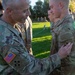 A senior U.S. Army soldier is pinning a unit patch onto the sleeve of a younger soldier during what appears to be a formal military ceremony outdoors. Both soldiers are wearing OCP (Operational Camouflage Pattern) uniforms, and the senior soldier has a 4th Infantry Division patch and U.S. flag on his left sleeve. Multiple other soldiers are visible in formation in the background on a green lawn.