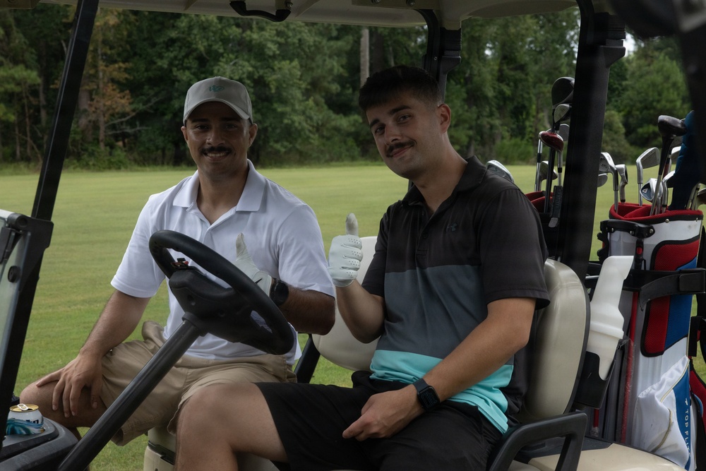 Two men are seated in a golf cart on what appears to be a golf course. The passenger on the right is giving a thumbs-up gesture toward the camera, while the driver on the left is smiling. Both men have mustaches and are dressed in golf attire.