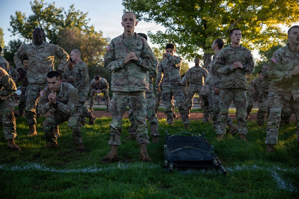 A group of U.S. Army soldiers in OCP (Operational Camouflage Pattern) uniforms are gathered on a grass field, appearing to be engaged in a physical training or drill session. The central figure, whose name tape reads 'CHETWOOD,' stands upright with arms crossed while others around him are in crouched or resting positions. The setting appears to be an outdoor military training area during early morning or evening light.