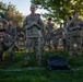 A group of U.S. Army soldiers in OCP (Operational Camouflage Pattern) uniforms are gathered on a grass field, appearing to be engaged in a physical training or drill session. The central figure, whose name tape reads 'CHETWOOD,' stands upright with arms crossed while others around him are in crouched or resting positions. The setting appears to be an outdoor military training area during early morning or evening light.