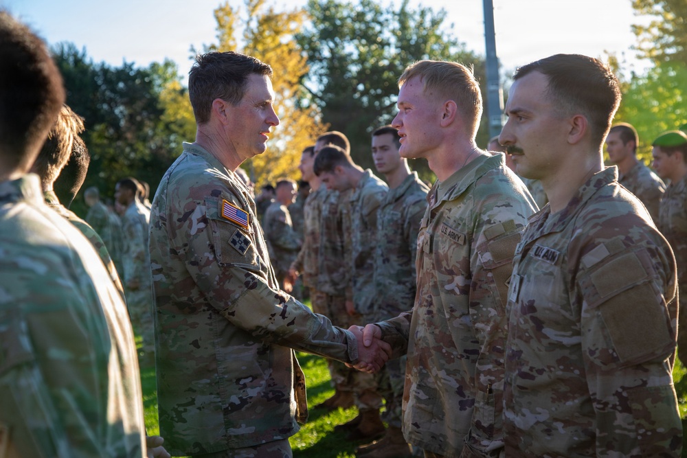 A senior U.S. Army officer is shaking hands with soldiers lined up in formation outdoors. The scene appears to be a formal recognition or greeting ceremony, with multiple soldiers in Operational Camouflage Pattern (OCP) uniforms standing at attention. The setting is a grassy outdoor area with trees visible in the background.