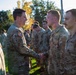 A senior U.S. Army officer is shaking hands with soldiers lined up in formation outdoors. The scene appears to be a formal recognition or greeting ceremony, with multiple soldiers in Operational Camouflage Pattern (OCP) uniforms standing at attention. The setting is a grassy outdoor area with trees visible in the background.