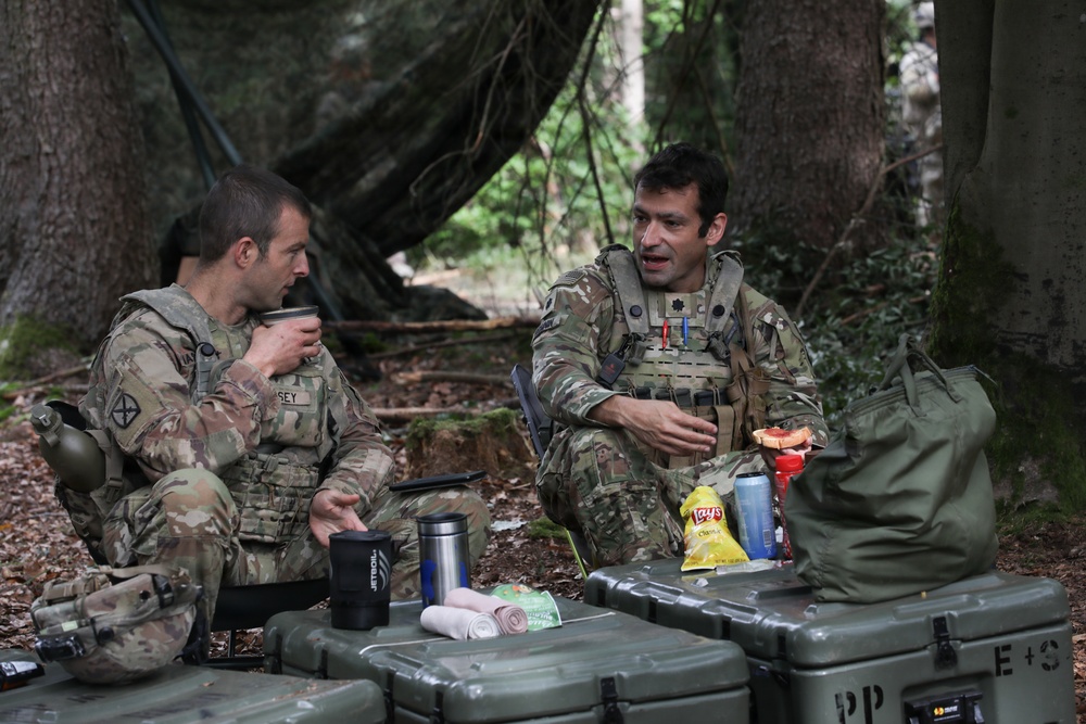 Two soldiers in multicam/OCP camouflage uniforms are taking a break in a forested area, seated on the ground near military equipment cases. They appear to be in conversation while eating and drinking, with snacks and beverages visible between them. The setting suggests a field exercise or operational pause in a wooded environment.