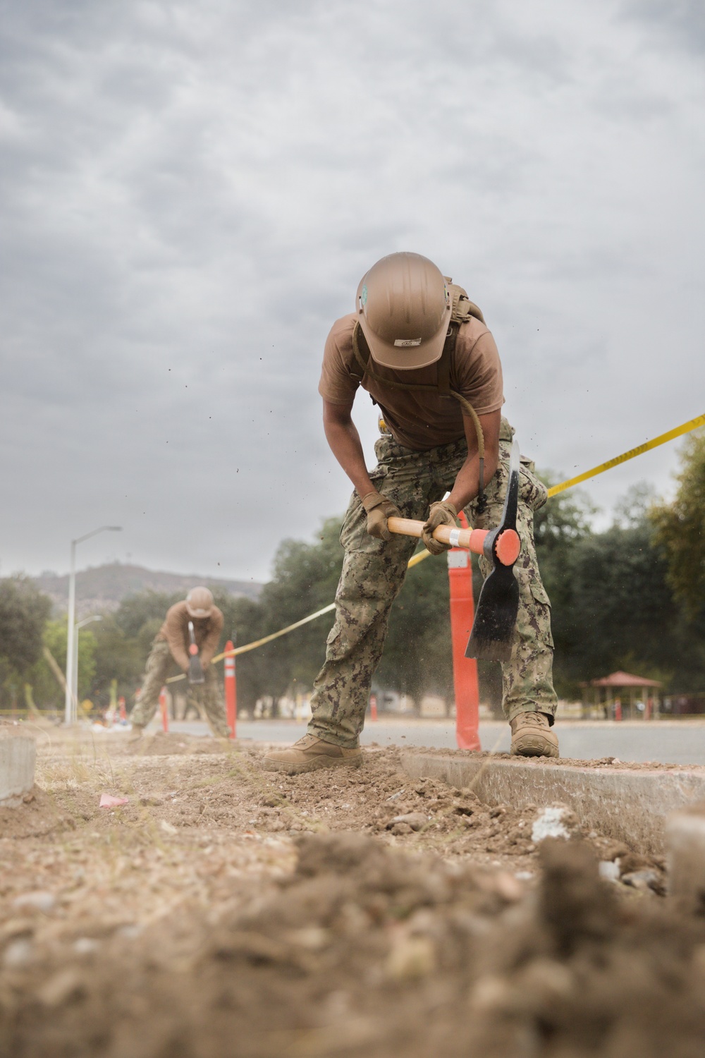 Two military personnel in camouflage uniforms and tan hard hats are performing construction or road work, using pickaxes to break up soil or pavement along a curbed roadway. The work area is cordoned off with orange traffic cones and yellow caution tape. The setting appears to be a military installation or base, with trees, a street lamp, and a gazebo visible in the background.