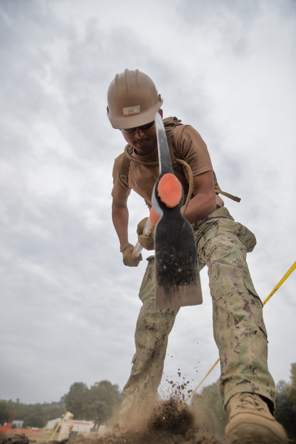 A military service member wearing a tan hard hat labeled 'CAO', safety glasses, and camouflage uniform is vigorously digging with a shovel at a construction or field site. The photo is taken from a dramatic low-angle perspective, with the shovel blade prominently in the foreground and dirt flying from the impact. The individual appears to be part of a military engineering or construction unit based on their gear and attire.