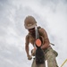 A military service member wearing a tan hard hat labeled 'CAO', safety glasses, and camouflage uniform is vigorously digging with a shovel at a construction or field site. The photo is taken from a dramatic low-angle perspective, with the shovel blade prominently in the foreground and dirt flying from the impact. The individual appears to be part of a military engineering or construction unit based on their gear and attire.