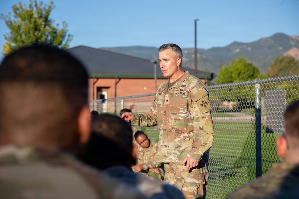 A U.S. Army officer in Operational Camouflage Pattern (OCP) uniform is addressing a group of soldiers outdoors on what appears to be a military installation. The officer appears to be speaking or briefing the assembled personnel, who are seated or standing with their backs to the camera. Mountains and brick buildings are visible in the background.