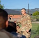 A U.S. Army officer in Operational Camouflage Pattern (OCP) uniform is addressing a group of soldiers outdoors on what appears to be a military installation. The officer appears to be speaking or briefing the assembled personnel, who are seated or standing with their backs to the camera. Mountains and brick buildings are visible in the background.