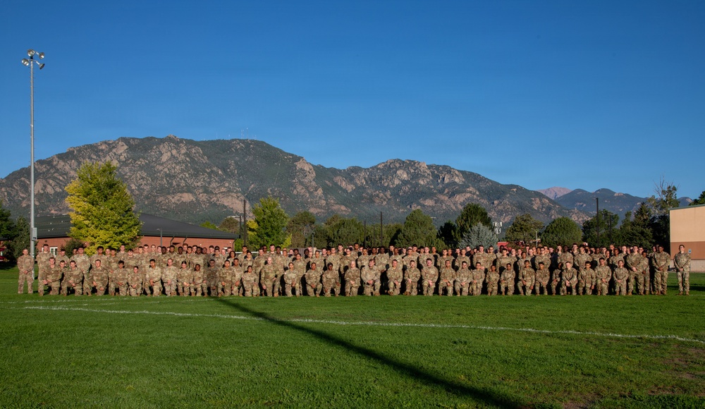 A large group of military personnel in camouflage uniforms are posed for a formal unit photograph on a grass athletic field. The backdrop features a prominent Rocky Mountain range under a clear blue sky, consistent with the Colorado Springs/Fort Carson area. The group consists of several rows of soldiers arranged in a wide formation spanning the width of the field.