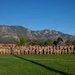 A large group of military personnel in camouflage uniforms are posed for a formal unit photograph on a grass athletic field. The backdrop features a prominent Rocky Mountain range under a clear blue sky, consistent with the Colorado Springs/Fort Carson area. The group consists of several rows of soldiers arranged in a wide formation spanning the width of the field.