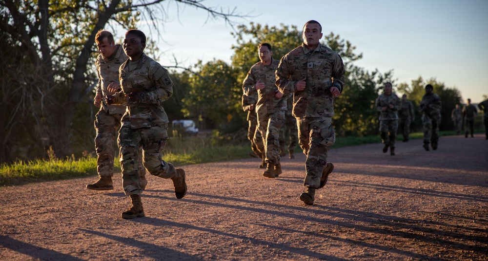 A group of military personnel in camouflage uniforms are running along a gravel path during what appears to be early morning or evening, based on the warm, low-angle lighting. The soldiers are wearing OCP (Operational Camouflage Pattern) uniforms consistent with U.S. Army or Air Force attire. This appears to be a physical training (PT) run with multiple service members participating.