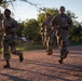 A group of military personnel in camouflage uniforms are running along a gravel path during what appears to be early morning or evening, based on the warm, low-angle lighting. The soldiers are wearing OCP (Operational Camouflage Pattern) uniforms consistent with U.S. Army or Air Force attire. This appears to be a physical training (PT) run with multiple service members participating.