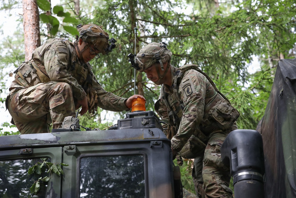 Two soldiers in U.S. Army MultiCam/OCP camouflage uniforms are working on top of a military vehicle, appearing to install or adjust an amber warning light. Both soldiers are wearing combat helmets equipped with night vision goggle mounts and full tactical gear. The setting is a wooded area, suggesting a field training exercise or operational environment.