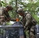 Two soldiers in U.S. Army MultiCam/OCP camouflage uniforms are working on top of a military vehicle, appearing to install or adjust an amber warning light. Both soldiers are wearing combat helmets equipped with night vision goggle mounts and full tactical gear. The setting is a wooded area, suggesting a field training exercise or operational environment.