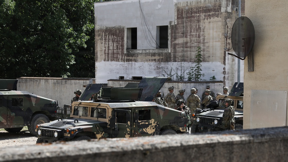 The image shows a group of military personnel gathered around multiple Humvee (HMMWV) vehicles in what appears to be an urban or semi-urban compound area. Soldiers in combat uniforms and helmets are clustered together, possibly conducting a briefing or vehicle maintenance check. The setting includes a weathered building with a satellite dish and surrounding vegetation.