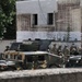 The image shows a group of military personnel gathered around multiple Humvee (HMMWV) vehicles in what appears to be an urban or semi-urban compound area. Soldiers in combat uniforms and helmets are clustered together, possibly conducting a briefing or vehicle maintenance check. The setting includes a weathered building with a satellite dish and surrounding vegetation.